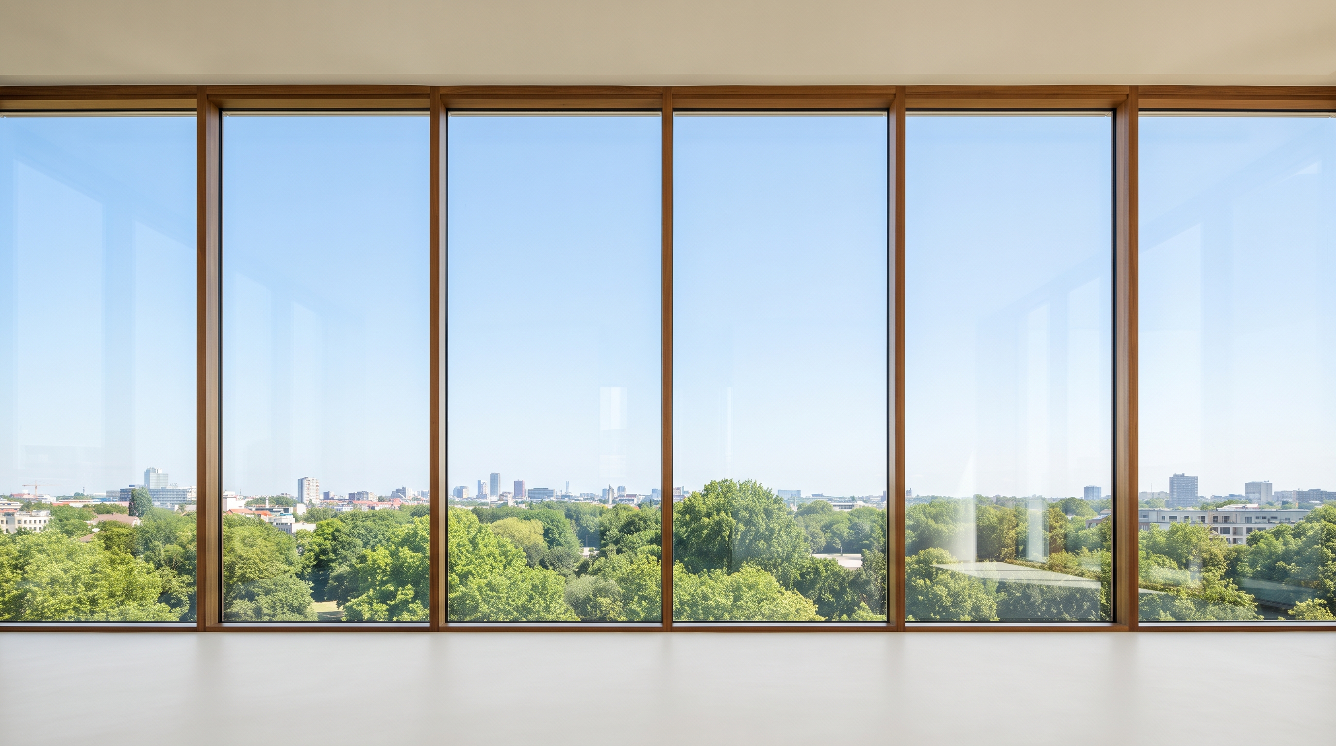 An empty minimal high-rise room with a flat floor-to-ceiling glass wall in warm wood mullions under a clear blue sky, green treetops and a low distant city skyline, pale matte floor.