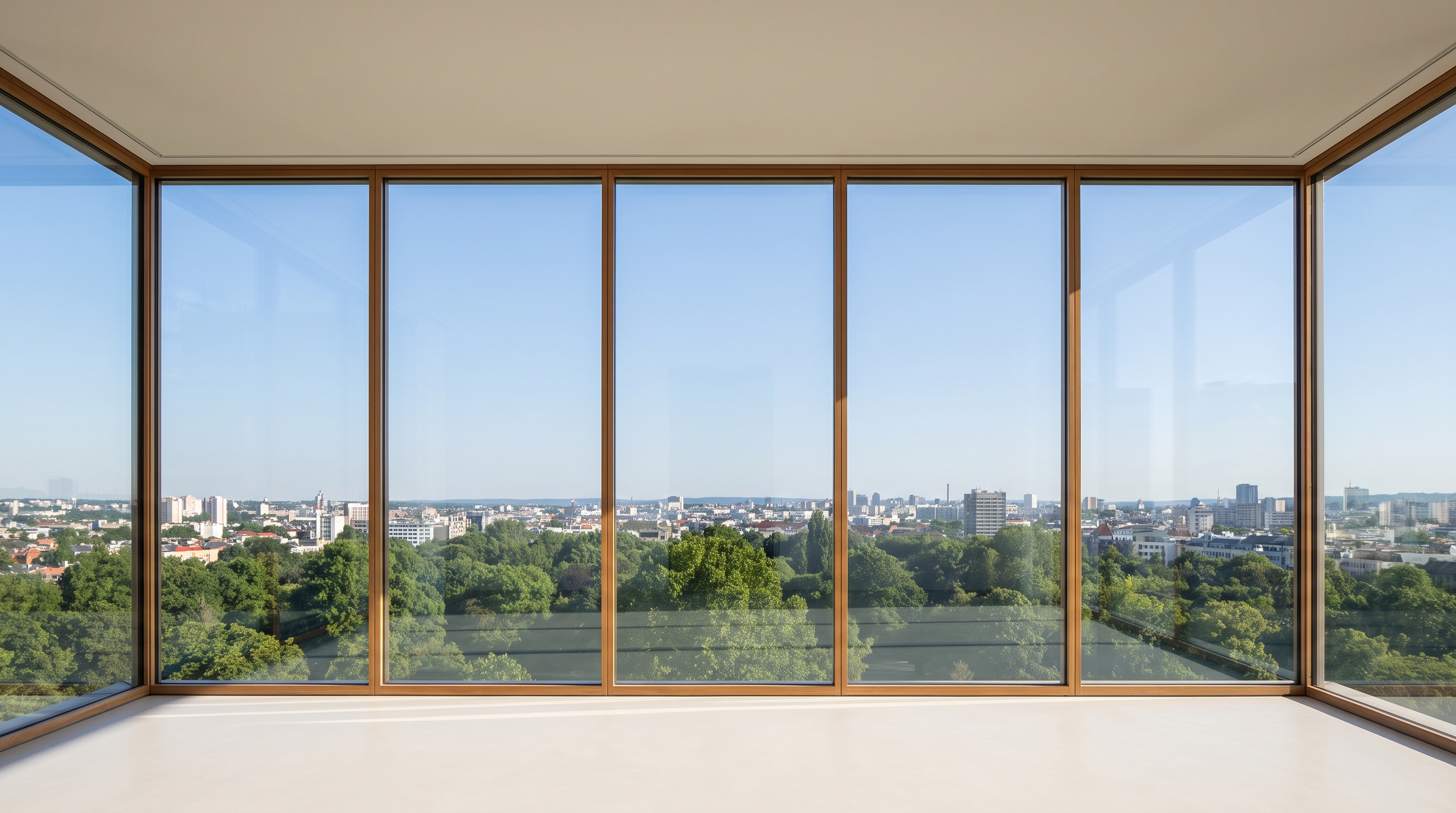 An empty minimal high-rise corner room with wood-framed glass wrapping the corner under a clear blue sky, green treetops and a distant city, pale matte floor.