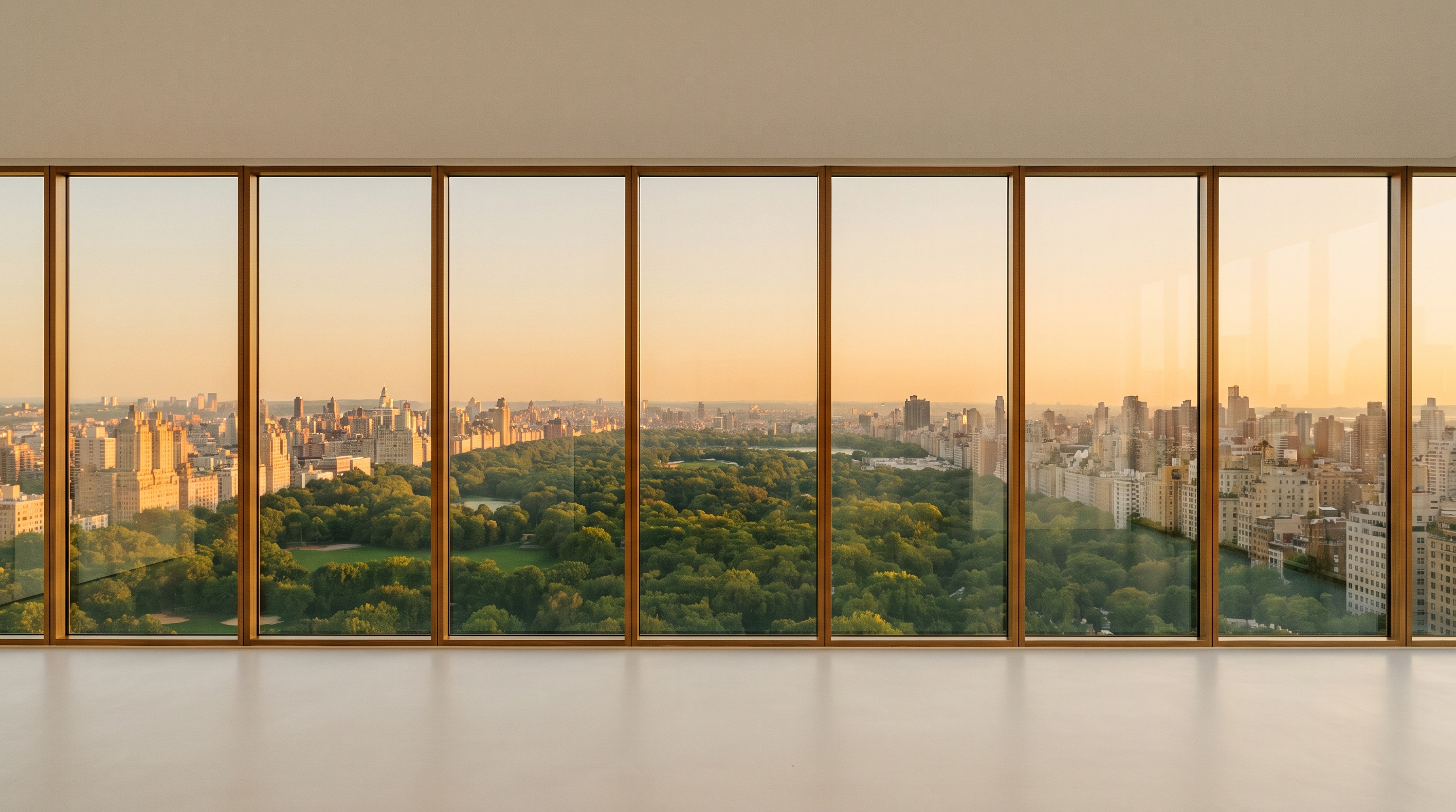 An empty minimal high-rise room with a flat floor-to-ceiling glass wall in wood mullions, a wide view over a green park and a dense glowing city skyline at golden hour, pale matte floor.