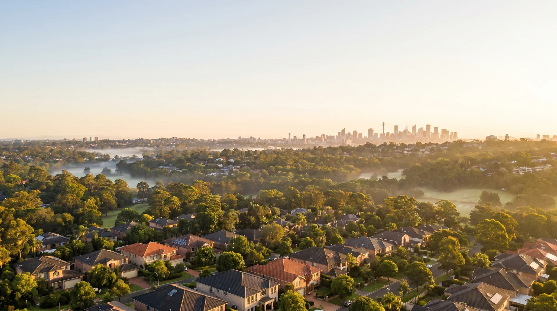 An elevated aerial over the Hills District at first light, soft mist settling between green canopy and rooftops, the distant Sydney skyline catching the morning sun.