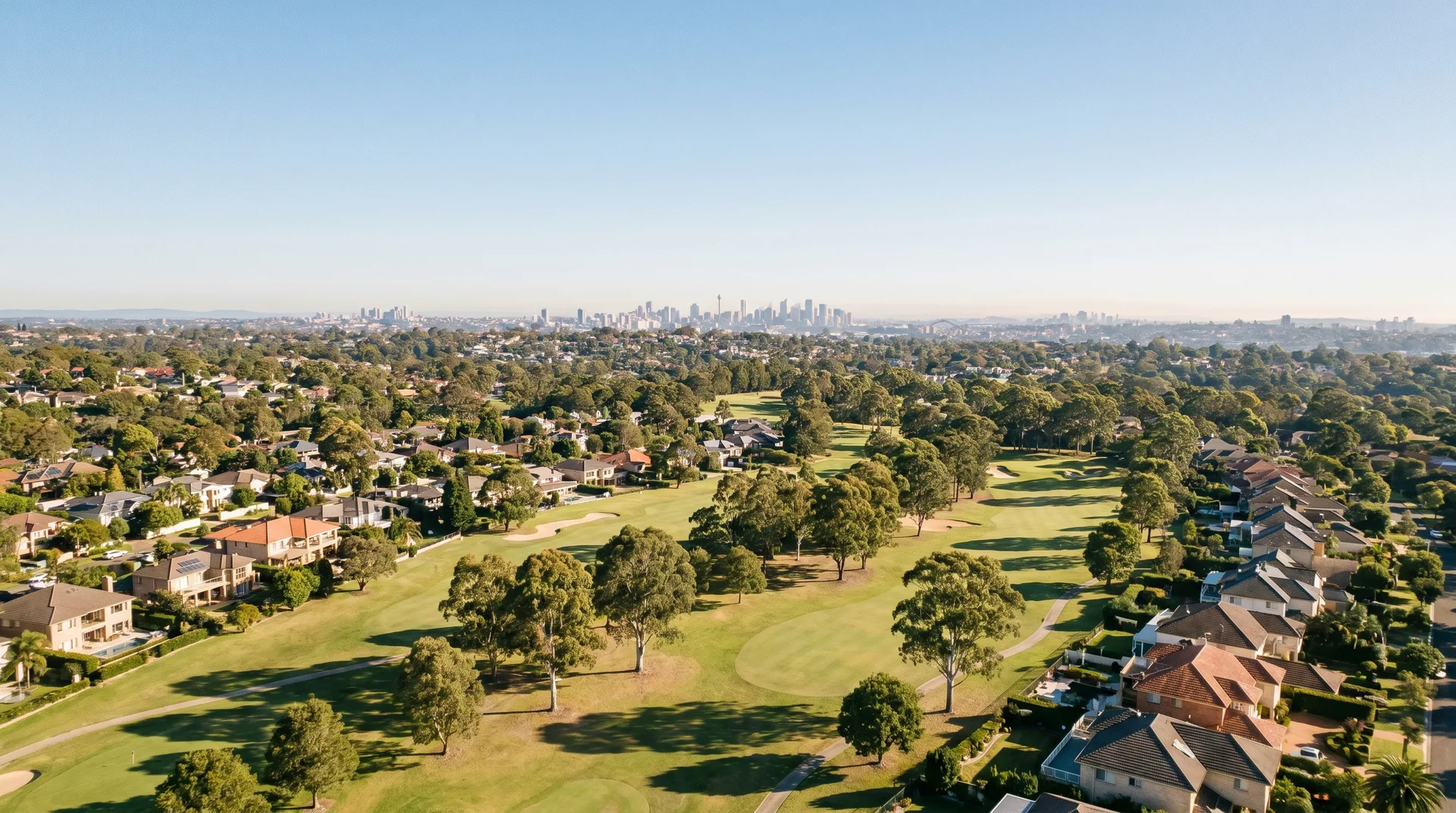 A high aerial over the Hills District showing a manicured golf course winding through green canopy and homes, with the distant Sydney skyline on the horizon.