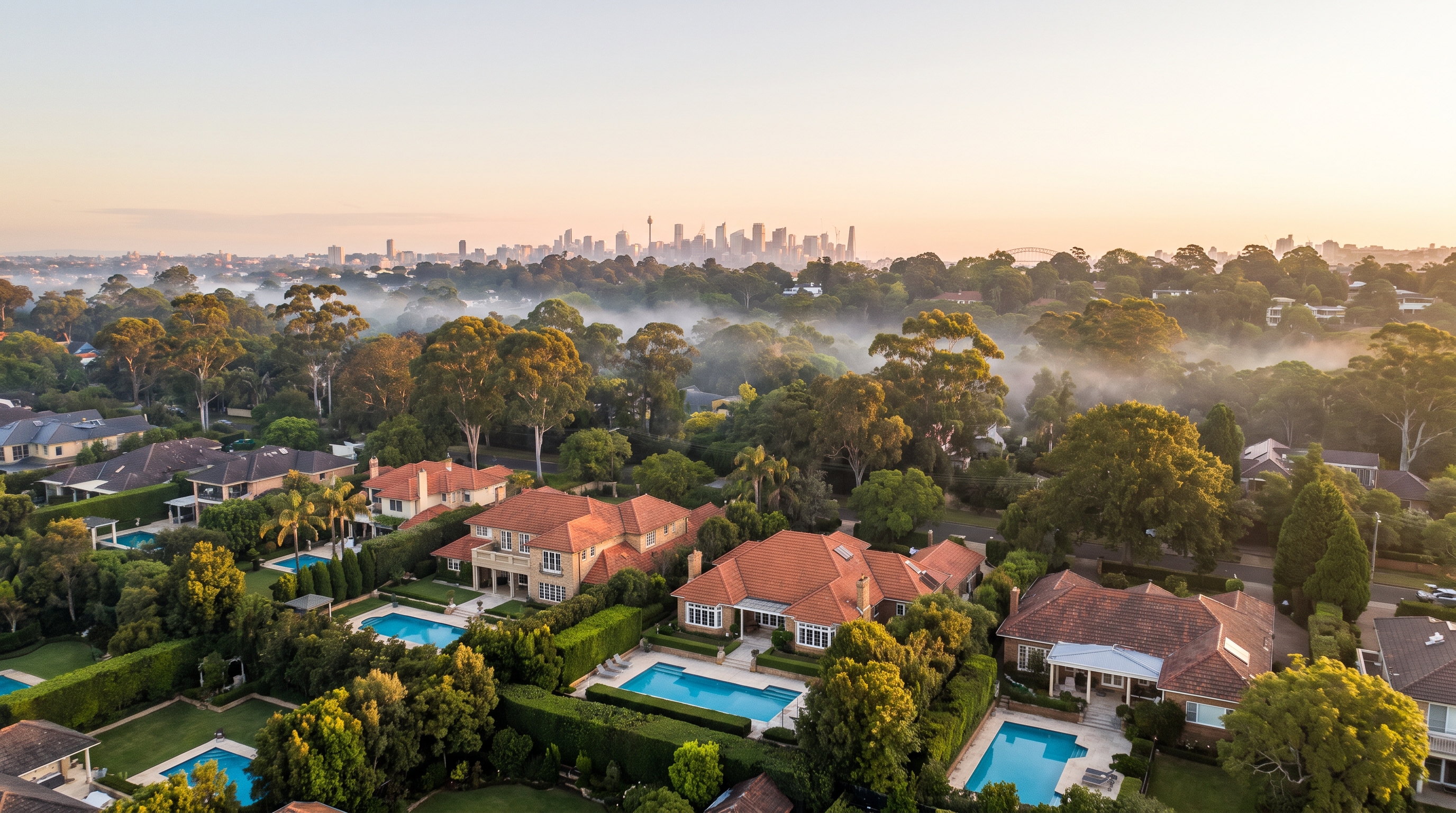 An aerial over substantial family homes with glittering pools and established gardens among warm green canopy in soft golden morning light, soft mist between the trees and the distant Sydney skyline on the horizon, an alternate framing.