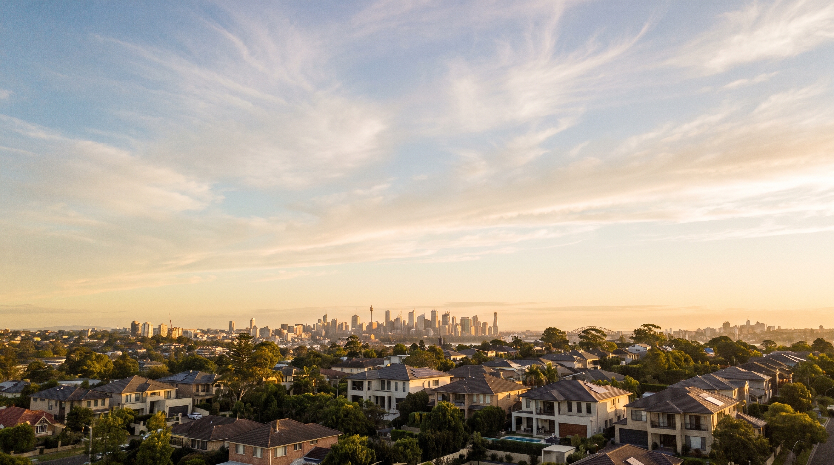An aerial at golden first light over large modern family homes and leafy gardens on a low horizon, the distant Sydney skyline glowing beyond, soft streaks of high cirrus cloud catching the warm light across the open upper sky.