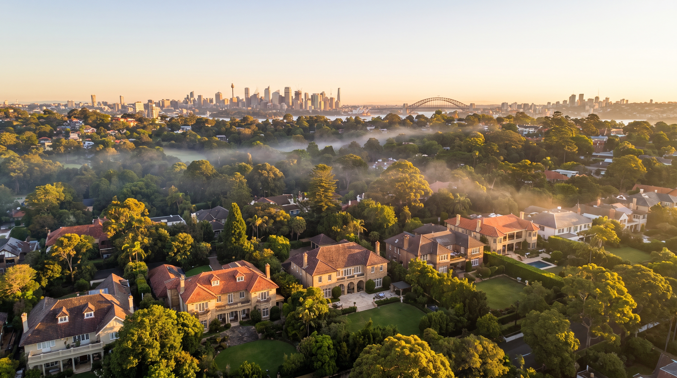 A high wide aerial over an affluent leafy suburb of large family homes and gardens glowing in warm morning light, gentle low mist threading the green canopy and the distant Sydney skyline warm on the far horizon.